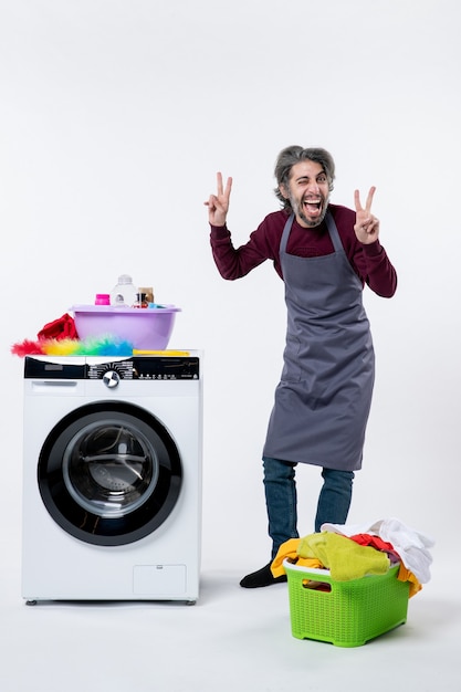 Vooraanzicht huishoudster man maken overwinning teken staande in de buurt van wasmachine wasmand op witte achtergrond