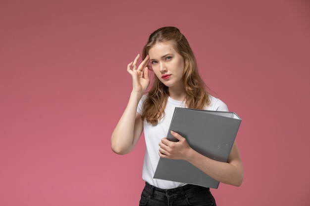 Vooraanzicht jong aantrekkelijk wijfje dat in wit t-shirt grijs dossier met denkende uitdrukking op het roze bureau houdt model vrouwelijk kleur vrouwelijk jong