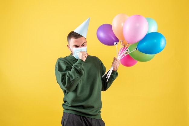 Vooraanzicht jong mannetje dat kleurrijke ballons in masker op gele achtergrond houdt