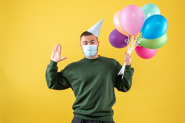 Vooraanzicht jong mannetje dat kleurrijke ballons in masker op gele achtergrond houdt