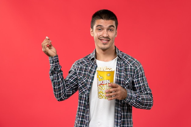 Vooraanzicht jong mannetje met popcornpakket met opgetogen uitdrukking op de lichtrode muur mannelijke bioscoop theater bioscoopfilm