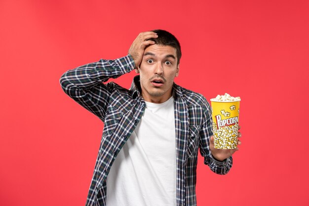 Vooraanzicht jong mannetje met popcornpakket met verbaasde uitdrukking op lichtrode muur mannelijke films theater bioscoopfilm