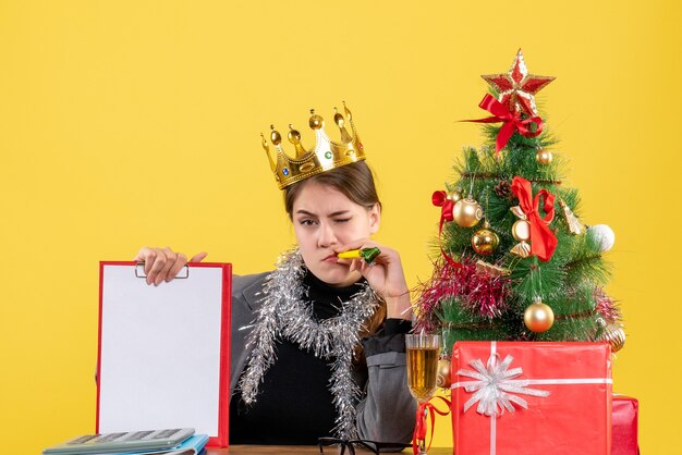 Vooraanzicht jong meisje met kroon zittend aan de tafel met document kerstboom en geschenken cocktail