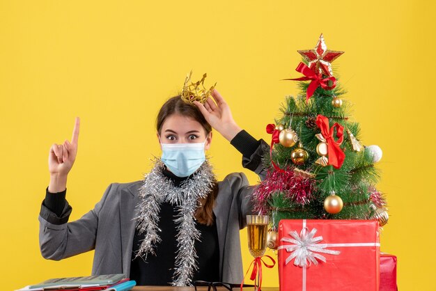 Vooraanzicht jong meisje met medisch masker dat kroon draagt die met vinger op Kerstmisboom en giftencocktail richt
