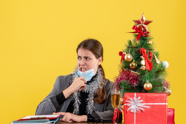 Vooraanzicht jong meisje met medisch masker zittend aan de tafel haar masker kerstboom en geschenken cocktail opstijgen