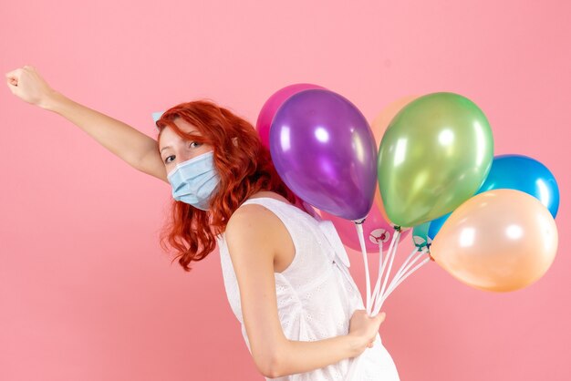 Vooraanzicht jong wijfje dat kleurrijke ballons in masker op roze bureau houdt van de pandemische covid-xmaspartij van het bureaukleurvirus