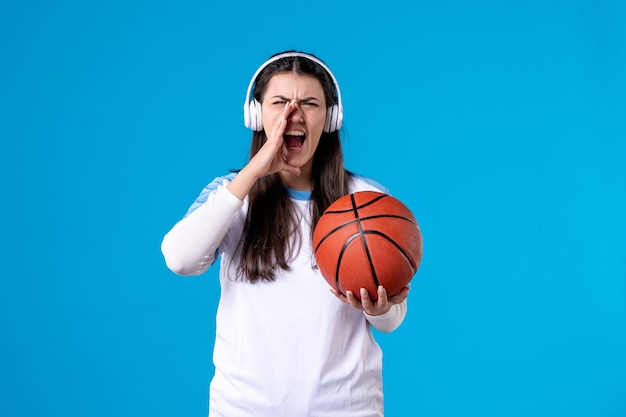 Vooraanzicht jong wijfje die met hoofdtelefoons basketbal op blauwe muur houden