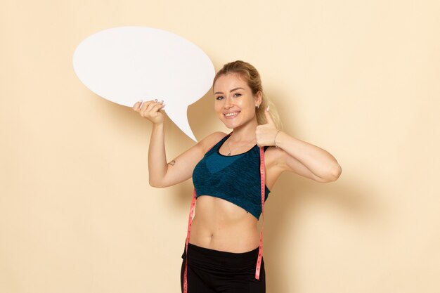 Vooraanzicht jong wijfje in sportuitrusting die wit bord houden en haar lichaam op een witte muur meten fit lichaam sport schoonheid gezondheid oefeningen