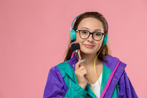 Gratis foto vooraanzicht jonge aantrekkelijke vrouw in wit t-shirt gekleurde jas luisteren naar muziek met koptelefoon op de roze achtergrond