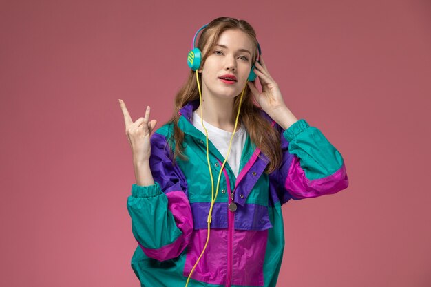 Vooraanzicht jonge aantrekkelijke vrouw in wit t-shirt gekleurde jas poseren luisteren naar muziek op het roze bureau model vrouw pose kleurenfoto