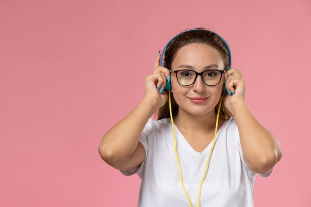 Vooraanzicht jonge aantrekkelijke vrouw in wit t-shirt gewoon poseren en luisteren naar muziek via koptelefoon lachend op de roze achtergrond