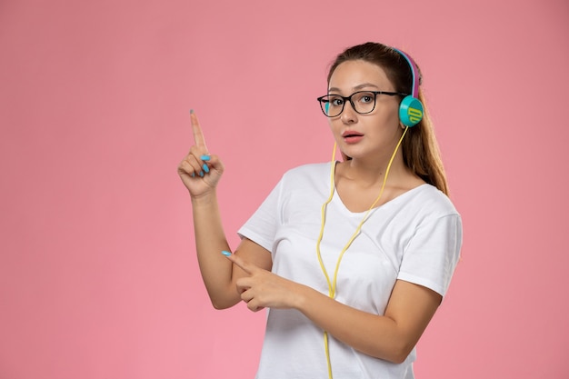 Vooraanzicht jonge aantrekkelijke vrouw in wit t-shirt luisteren naar muziek met koptelefoon op de roze achtergrond