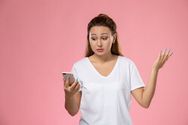 Vooraanzicht jonge aantrekkelijke vrouw in wit t-shirt met haar telefoon op roze achtergrond