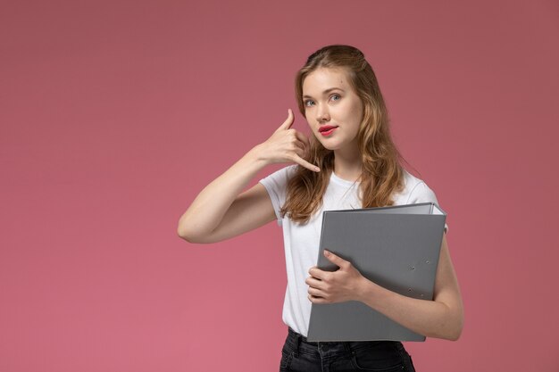 Vooraanzicht jonge aantrekkelijke vrouw in wit t-shirt poseren en houden grijs bestand op roze muur model vrouw pose kleurenfoto vrouwelijke jong