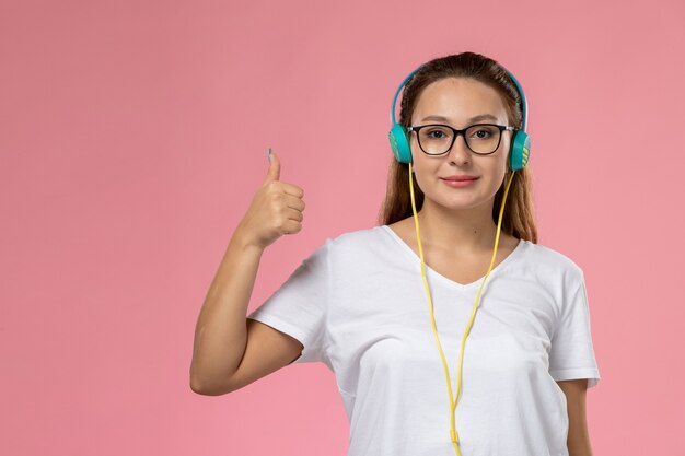 Vooraanzicht jonge aantrekkelijke vrouw in wit t-shirt poseren luisteren naar muziek met koptelefoon smi op de roze achtergrond