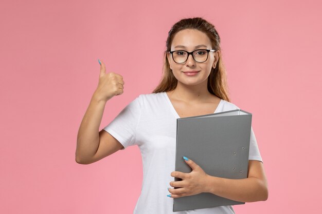 Vooraanzicht jonge aantrekkelijke vrouw in wit t-shirt poseren met grijs bestand smi op de roze achtergrond