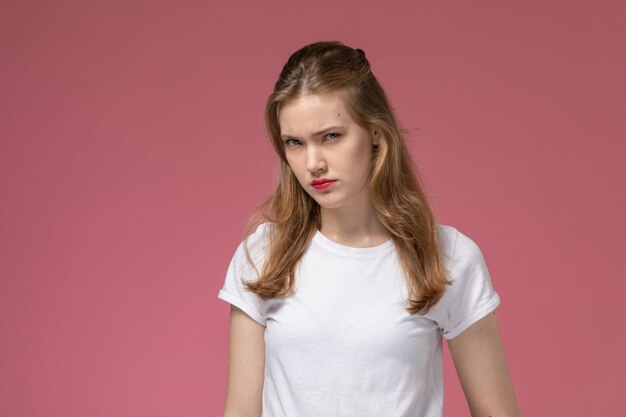 Vooraanzicht jonge aantrekkelijke vrouw in wit t-shirt poseren met ontevreden uitdrukking op roze muur model vrouw pose kleurenfoto