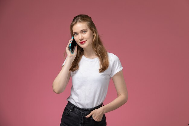 Vooraanzicht jonge aantrekkelijke vrouw in wit t-shirt praten aan de telefoon op roze muur model vrouwelijke pose kleurenfoto vrouwelijke jong