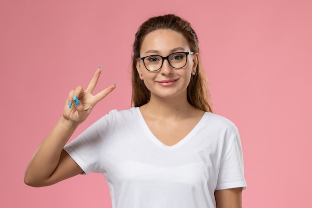 Vooraanzicht jonge aantrekkelijke vrouw in wit t-shirt smi en overwinningsteken waarop de roze achtergrond