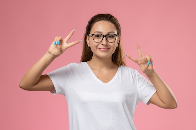 Vooraanzicht jonge aantrekkelijke vrouw in wit t-shirt smi met opgeheven overwinning gebaar vingers op de roze achtergrond