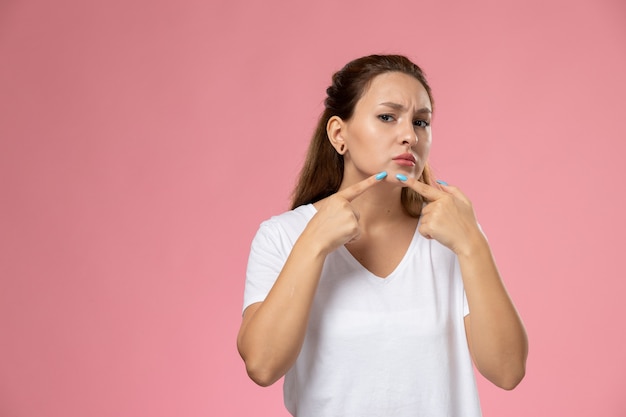 Vooraanzicht jonge aantrekkelijke vrouw in wit t-shirt wat betreft haar gezicht op de roze achtergrond