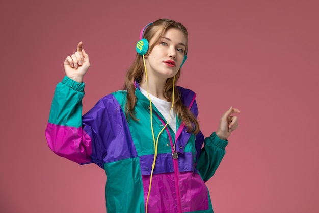 Vooraanzicht jonge aantrekkelijke vrouw luisteren naar muziek in gekleurde jas met dansbewegingen op de roze muur model kleur vrouwelijk jong meisje