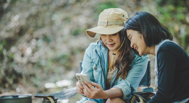 Vooraanzicht Jonge Aziatische mooie vrouw en haar vriendin zitten aan de voorkant van de tent gebruiken mobiele telefoon om foto te maken tijdens het kamperen in het bos met geluk samen