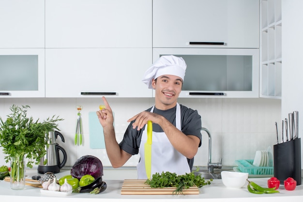 Vooraanzicht jonge chef-kok in uniform wijzend naar plafond in moderne keuken