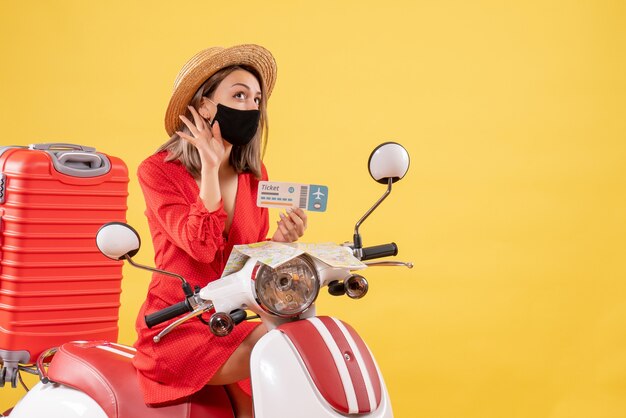 Vooraanzicht jonge dame op bromfiets met rode koffer met ticket luisterend naar iets