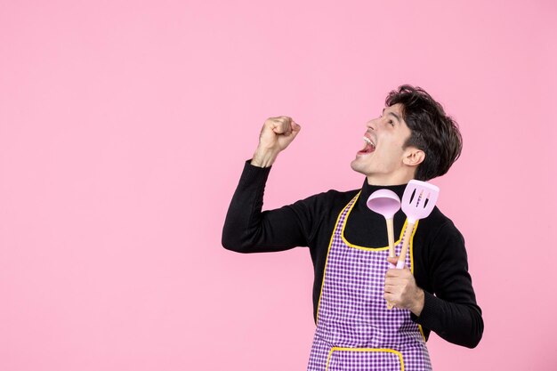 Vooraanzicht jonge man in cape die lepels vasthoudt en boven kijkt op roze achtergrond deeg beroep keuken uniform horizontale chef koken baan werknemer