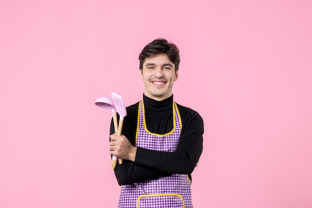 Vooraanzicht jonge man in cape met roze lepels op roze achtergrond beroep voedsel horizontale uniforme keuken kleur koken maaltijd deeg