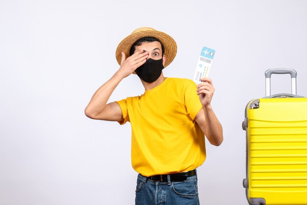 Vooraanzicht jonge man in geel t-shirt staande in de buurt van gele koffer reisticket bedekken oog met hand te houden