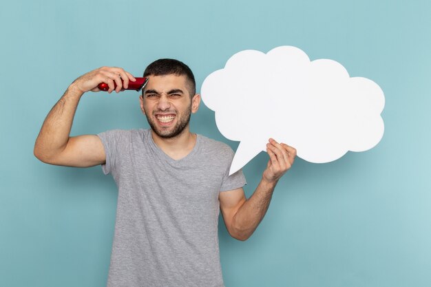 Vooraanzicht jonge man in grijs t-shirt met een groot wit bord en elektrisch scheerapparaat scheren op ijsblauwe muur mannelijke baard haar scheermes scheren