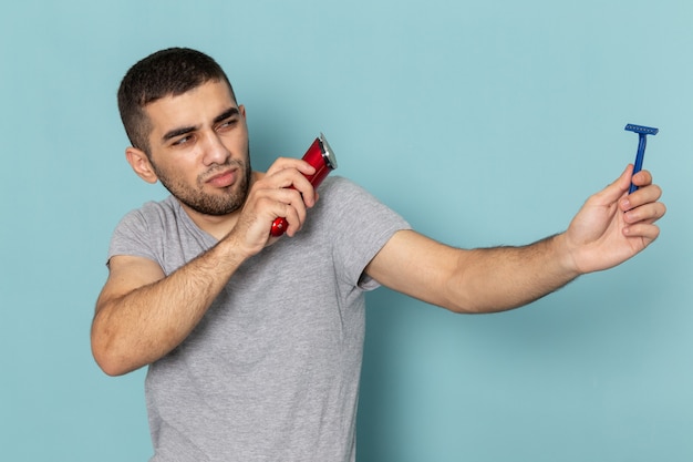 Vooraanzicht jonge man in grijs t-shirt met rood elektrisch scheerapparaat en eenvoudig scheerapparaat