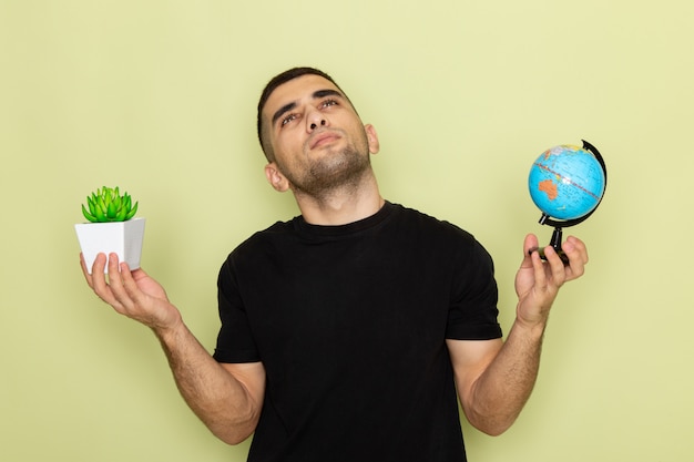 Vooraanzicht jonge man in zwart t-shirt met kleine groene plant en een kleine wereldbol terwijl hij aan groen denkt