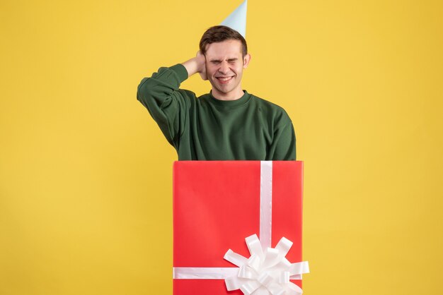 Vooraanzicht jonge man met feestmuts met zijn oren achter grote geschenkdoos op gele achtergrond