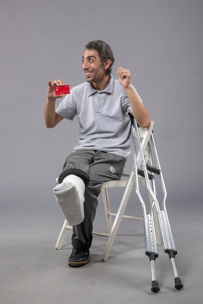 Vooraanzicht jonge man met gebroken voet en verband met bankkaart op de grijze muur ongeval twist menselijke pijn mannelijke voet
