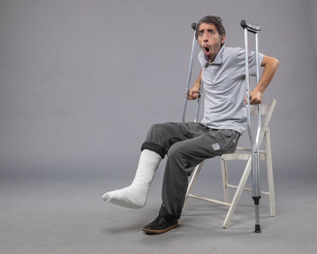 Vooraanzicht jonge man met gebroken voet probeert op te staan met krukken op de grijze muur voet gebroken pijn draai ongeval been