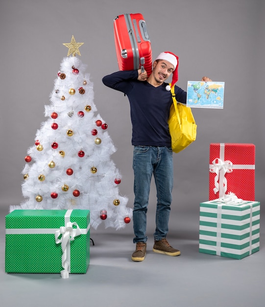 Vooraanzicht jonge man met gele rugzak met kaart en rode reistas in de buurt van kerstboom en cadeautjes op grijs geïsoleerd
