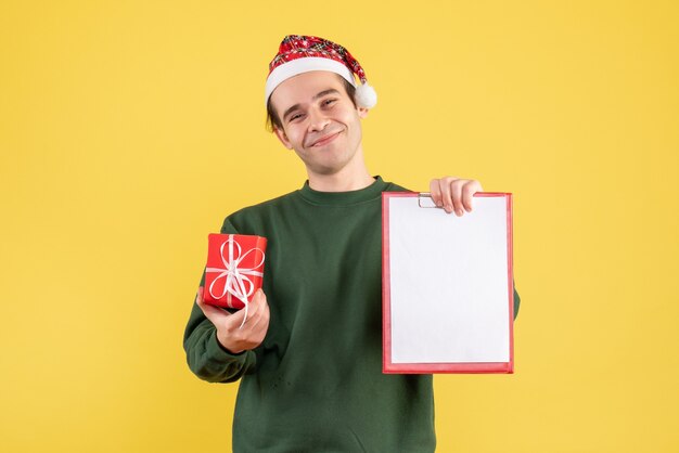 Vooraanzicht jonge man met groene trui met klembord en cadeau staande op geel