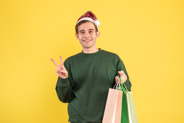 Vooraanzicht jonge man met kerstmuts met boodschappentassen overwinning teken maken op gele achtergrond