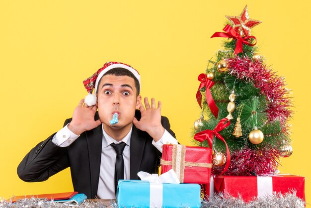 Vooraanzicht jonge man met kerstmuts zitten aan de tafel met behulp van noisemaker kerstboom en geschenken