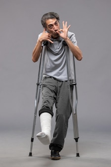 Vooraanzicht jonge man met krukken als gevolg van gebroken voet op de grijze muur pijn voet uitschakelen ongeval been gebroken