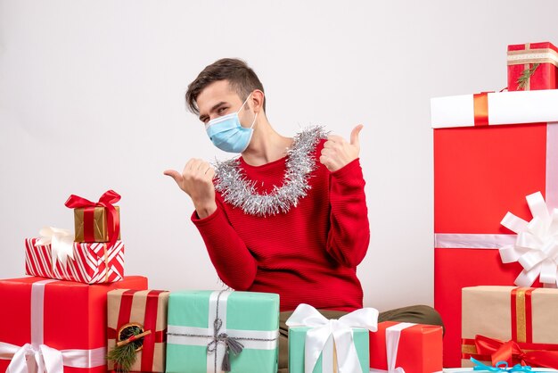 Vooraanzicht jonge man met masker duim omhoog teken met beide handen rond kerstcadeaus zitten