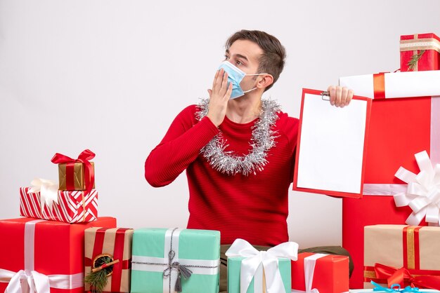 Vooraanzicht jonge man met masker houden klembord zittend op de vloer rond Kerstcadeaus