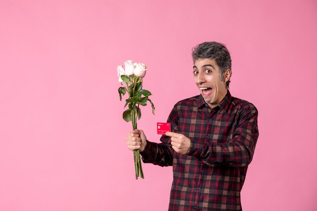 Vooraanzicht jonge man met mooie roze rozen en bankkaart op roze muur