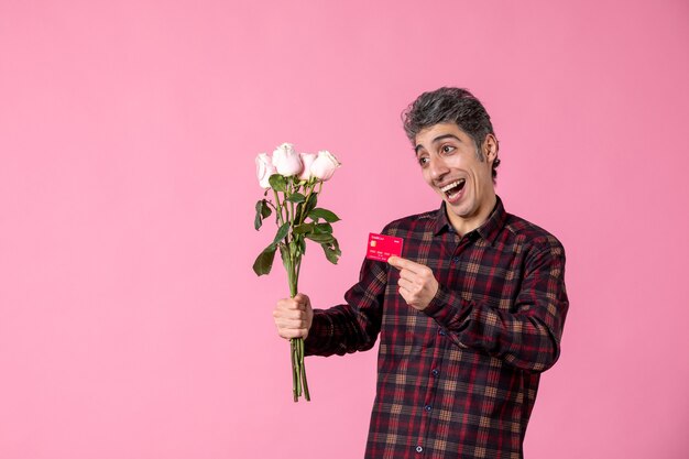 Vooraanzicht jonge man met mooie roze rozen en bankkaart op roze muur