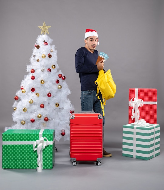 Vooraanzicht jonge man met reiskaartje en rode koffer die zijn gele rugzak controleert rond verschillende cadeautjes op grijs geïsoleerd