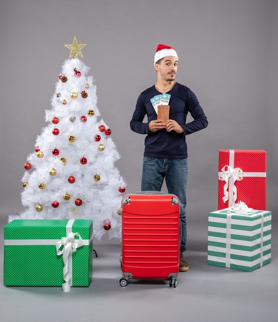 Vooraanzicht jonge man met rode koffer die zijn reiskaartjes met beide handen op grijs houdt