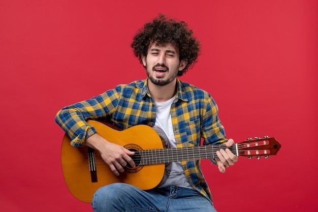 Vooraanzicht jonge man zit en speelt gitaar op de rode muur live concertmuzikant applaus band speelt muziek